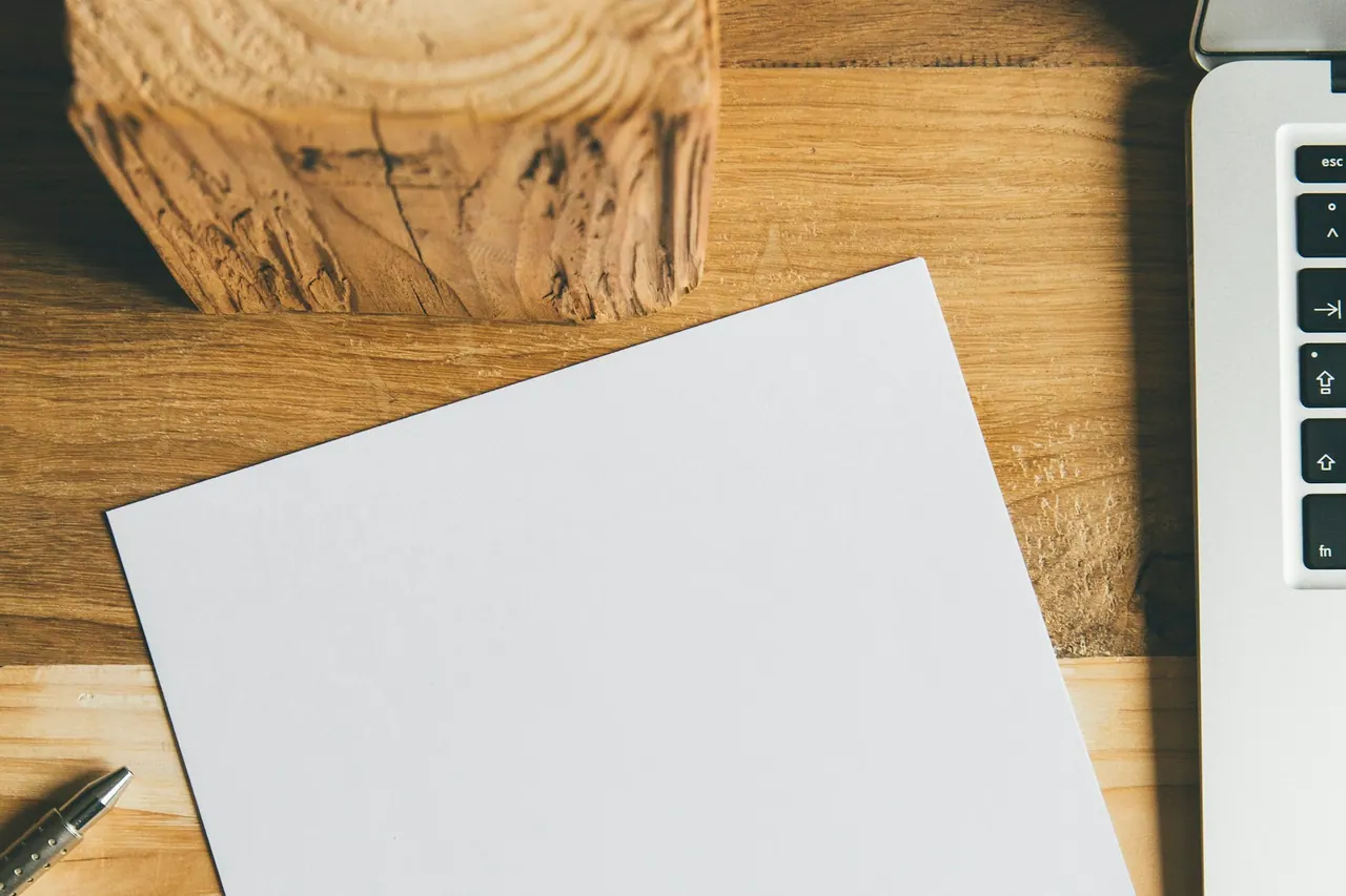 A blank sheet of paper and a pen beside a laptop on a wooden desk.
